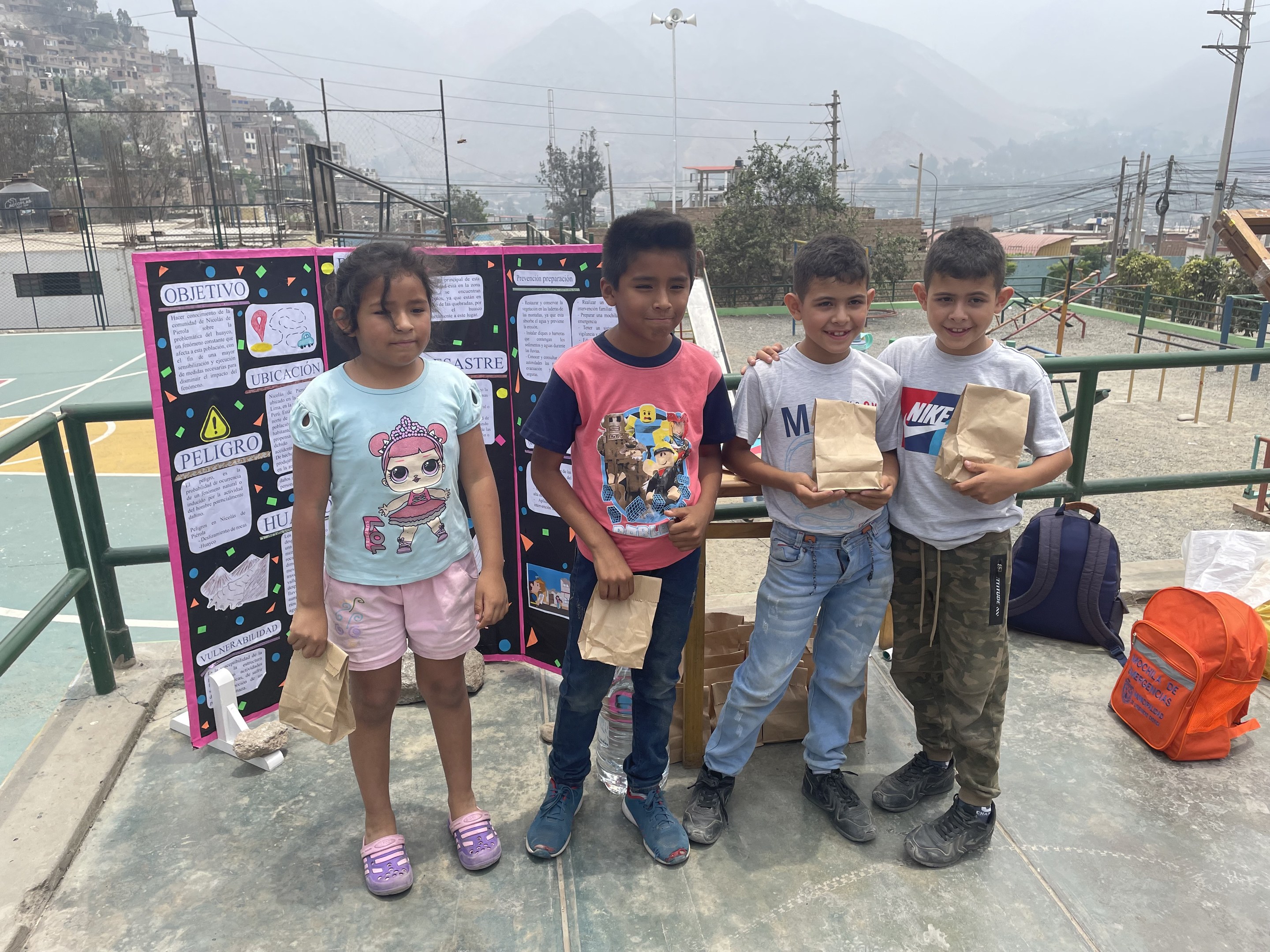 Gruppe von Kindern auf einem Basketballfeld mit Papierbeuteln in der Hand, mit Beuteln und Gegenständen rechts und einer Tafel mit Text links, vor einer Außenanlage mit Zaun, Pfählen, Straßenlaternen, Ästen, Gebäuden, Hügeln und Himmel.