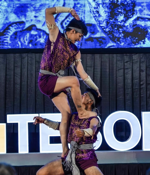 Zwei Akrobaten führen auf der Bühne eine Nummer auf, wobei der Mann die Frau in seinen Armen hält, während beide purpurne Kleider tragen, mit einem schwarzen Vorhang und einer Leinwand im Hintergrund und sichtbaren Zuschauern unten.