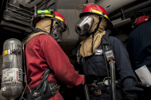 Zwei Feuerwehrleute in Helmen und Gasmasken neben einem Sauerstoffzylinder mit Rohren und anderen Gegenständen im Hintergrund.
