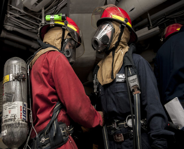Zwei Feuerwehrleute in Helmen und Gasmasken neben einem Sauerstoffzylinder mit Rohren und anderen Gegenständen im Hintergrund.