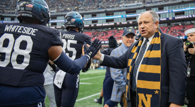 Ein Mann im Anzug schüttelt einem Fussballspieler auf einem Feld die Hand, umgeben von einer Menge, während eine Person mit einer Kamera und einer Mütze rechts steht; ein Stadion mit Zuschauern und Schildern, auf denen "Notre Dame Football Coach Mike McCarthy und die Fighting Irish" zu lesen sind, ist im Hintergrund zu sehen.