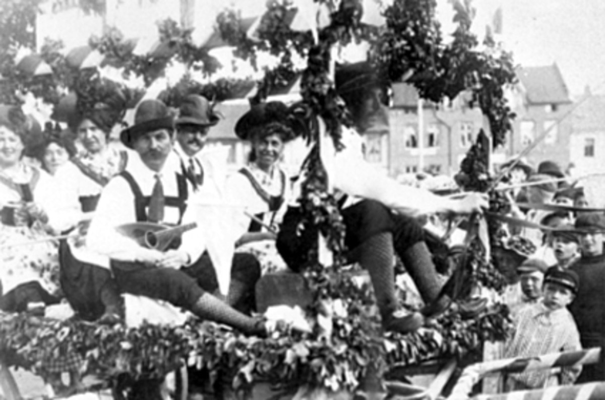 Schwarzes und weißes Bild eines Paradewagens, der Menschen in traditioneller bayrischer Tracht trägt, einige spielen Musikinstrumente, der Wagen ist mit Blättern und anderen Verzierungen geschmückt, vor einem Gebäude unter einem klaren Himmel.