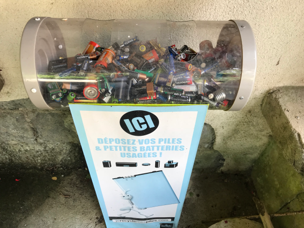 Ein Müllcontainer voller Batterien auf einem Gehweg, mit einem Plakat mit Text an der Wand dahinter und einer Treppe auf der rechten Seite.
