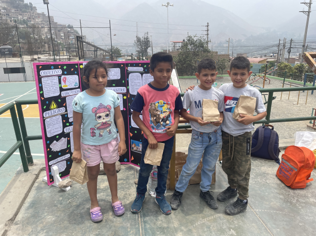 Gruppe von Kindern, die Papiertüten auf einem Basketballfeld halten, mit Tüten und Gegenständen auf dem Boden, einer Tafel mit Text links daneben und einem Zaun, Pfosten, Straßenlaternen, Bäumen, Gebäuden, Hügeln und Himmel im Hintergrund.