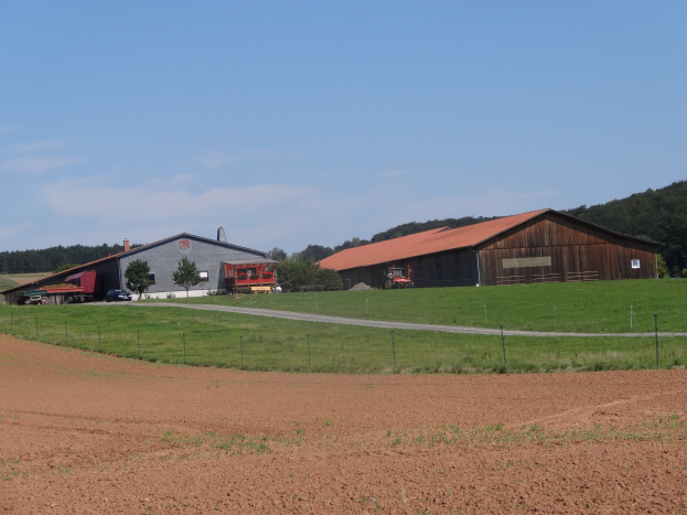 Ein Hof mit einem großen Stall in einer grünen Wiese, die von einem Zaun umgeben ist, ein paar geparkte Fahrzeuge davor, Bäume im Hintergrund und eine bewölkte Himmel.