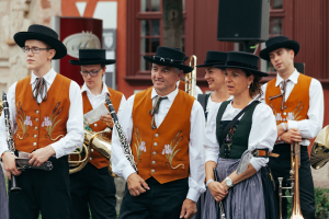 Gruppe von Menschen in traditioneller bayrischer Tracht mit Musikinstrumenten, die neben einem Gebäude mit Fenstern, einem Lautsprecher und Pflanzen im Hintergrund stehen.