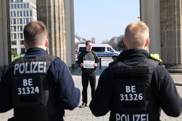 Drei Polizeibeamte stehen vor einem Gebäude, ein Mann in der Mitte hält ein Papier, flankiert von Säulen; Fahrzeuge, Menschen, Bäume, Gebäude und der Himmel sind im Hintergrund sichtbar.