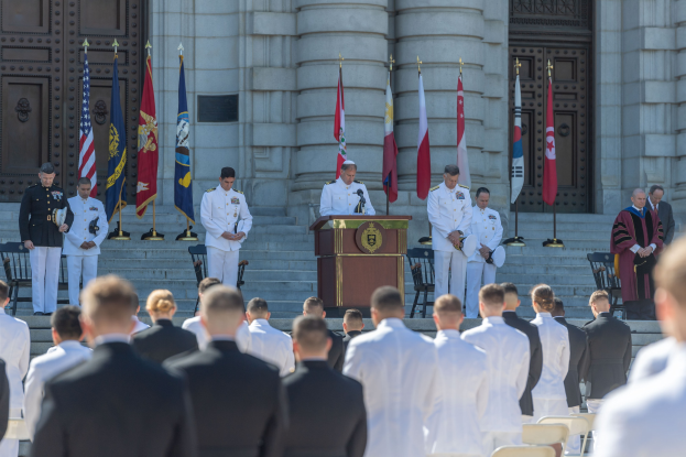 Gruppe von Männern in weißer Uniformen vor einem Gebäude mit Säulen und Türen, mit einem Podium und Mikrofon in der Mitte, Fahnen im Hintergrund und Stufen, die zum Gebäude führen.