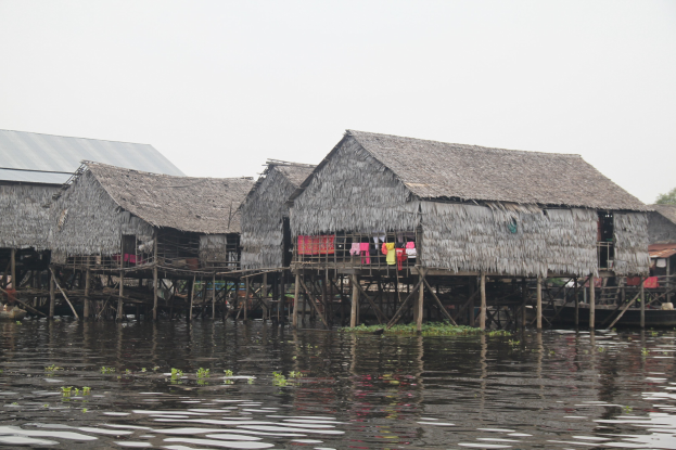 Eine Gruppe von Hütten auf einem Gewässer mit zum Trocknen aufgehängter Kleidung auf Holzpfählen, Bäumen und einem klaren blauen Himmel im Hintergrund.