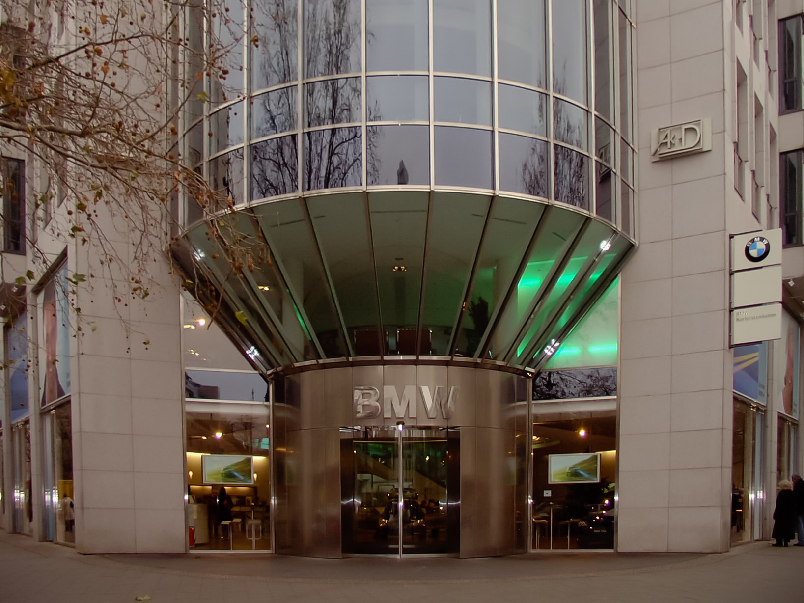 Großes Glasgebäude der BMW-Zentrale in Frankfurt, Deutschland, mit Text an der Fassade, Menschen davor und einem Baum links.