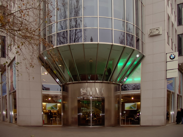 Großes Glasgebäude der BMW-Zentrale in Frankfurt, Deutschland, mit Text an der Fassade, Menschen davor und einem Baum links.