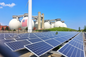 Solarpanels auf dem Dach eines Gebäudes mit einer Flagge, umgeben von einem Zaun, Gras, Pflanzen, Bäumen, Pfählen und anderen Gegenständen, mit Gebäuden und einem bewölkten Himmel im Hintergrund.