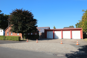 Straße mit Verkehrskegeln, die zu einem Gebäude mit Solarpanelen auf dem Dach führen, umgeben von Bäumen und Pflanzen unter einem klaren blauen Himmel.