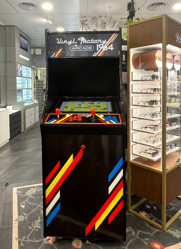 Ein Geschäft mit einem zentralen Vintage-Arcade-Automaten, einem Gestell mit Gläsern auf der rechten Seite und Hintergrund-Elementen wie Schränken, einem Fernseher und anderen Gegenständen, mit Deckenleuchten, die eine Retro-Atmosphäre schaffen.