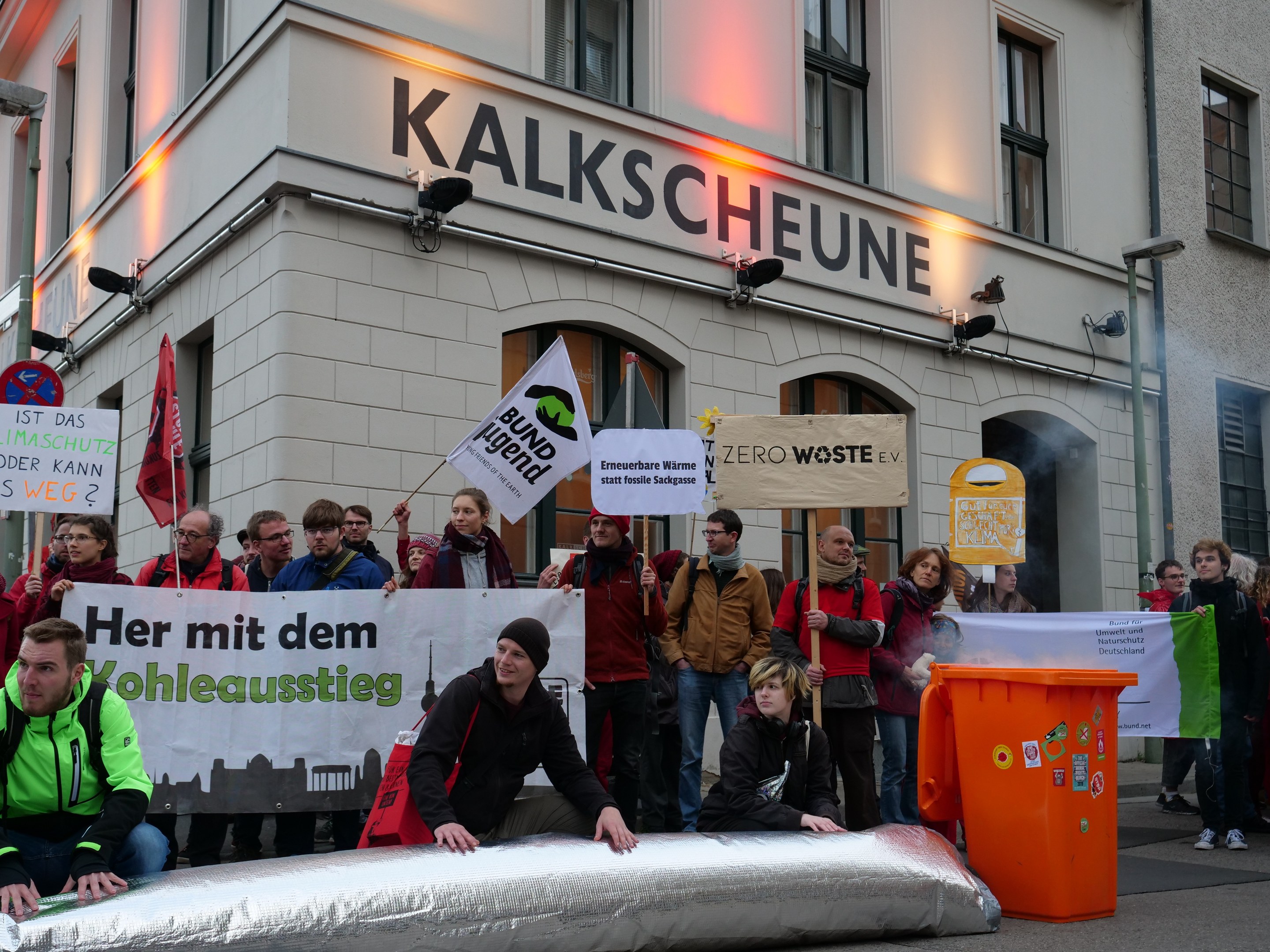 Eine Gruppe von Menschen mit Protestschildern und Transparenten vor einem Gebäude, mit zwei Personen im Vordergrund und einem Müllcontainer auf der rechten Seite.