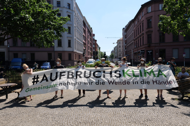 Menschen mit Masken halten ein Banner mit der Aufschrift 'Aufbruchsklima' während einer Klimaprotest in Berlin, umgeben von städtischen Elementen und anderen Teilnehmern.