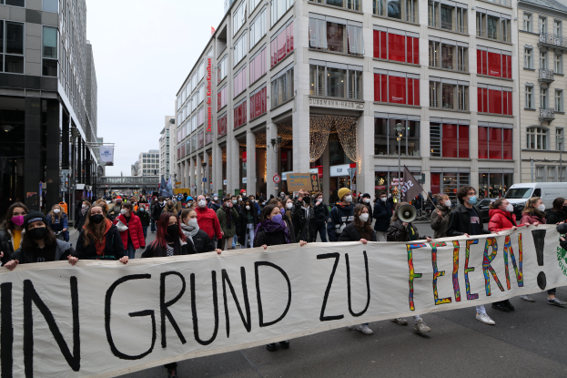 Eine Gruppe maskierter Personen hält ein Transparent mit der Aufschrift 'In Grund zu Feiern' vor Gebäuden, Laternenmasten und Fahrzeugen, was auf eine Protestaktion in Berlin, Deutschland, hindeutet.