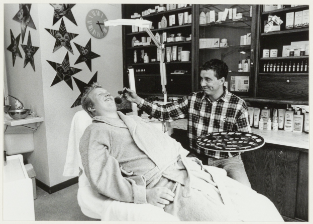 Ein Mann lächelt in einem Zahnarztstuhl, während ein anderer Mann neben ihm steht und ein Tablett hält, mit einem Waschbecken, Sternen, einer Uhr und Regalen voller Flaschen im Hintergrund.